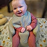 baby, infant, smiling, happy, pink_clothing, bib, baby_seat, tablecloth, animal_print, indoor, portrait, child, cute, hands, face, head, sitting, young_child, soft_lighting, blurred_background