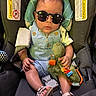 baby, child, car_seat, sunglasses, plush_toy, dinosaur_toy, clothing, footwear, necklace, person, infant, seatbelt, accessory, cute, fashion, portrait, indoor, headrest, sitting, adorable