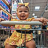 baby, child, infant, smiling, happy, shopping_cart, supermarket, grocery_store, yellow_dress, headband, bow, portrait, sitting, indoor, retail, diaper_aisle, cart_handle, hands, family_outing, colorful