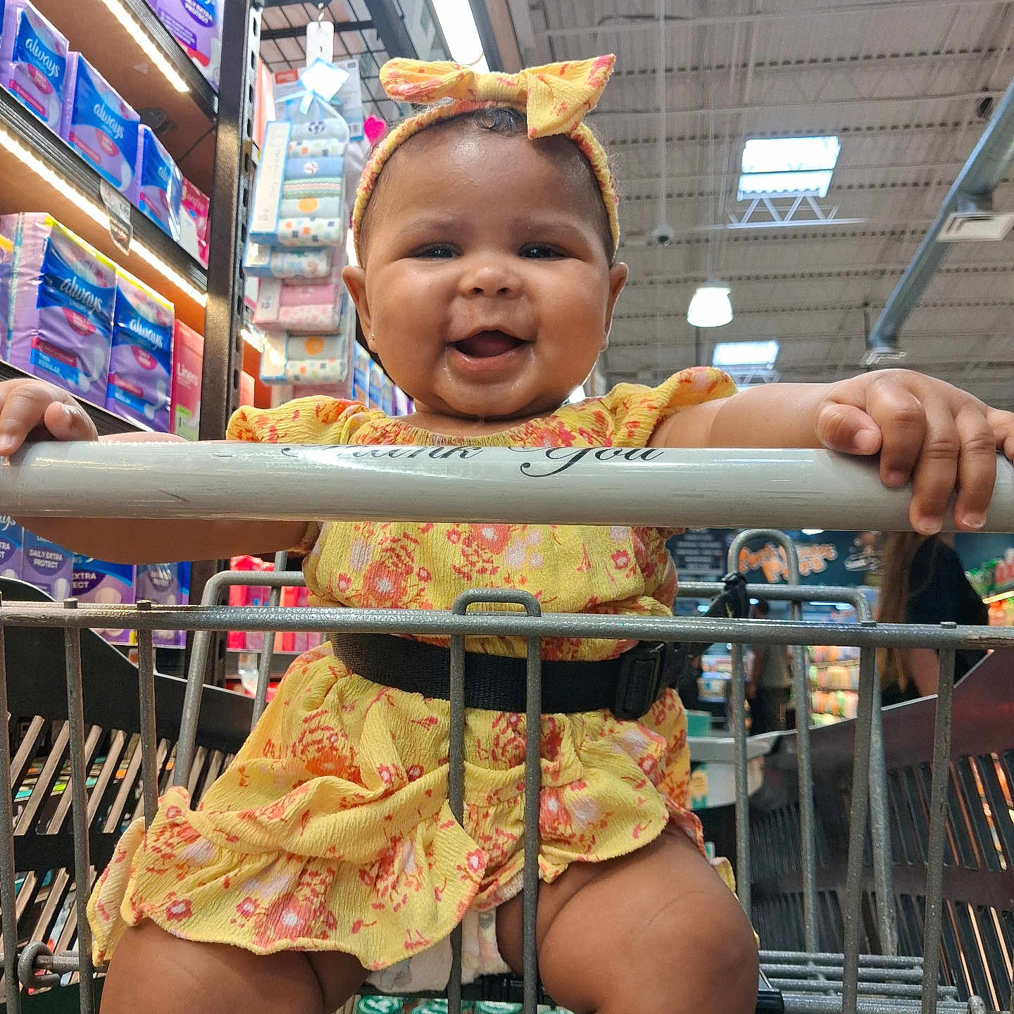 Ny'Linee is registered to the contest to win money with this photo: baby, child, infant, smiling, happy, shopping_cart, supermarket, grocery_store, yellow_dress, headband, bow, portrait, sitting, indoor, retail, diaper_aisle, cart_handle, hands, family_outing, colorful