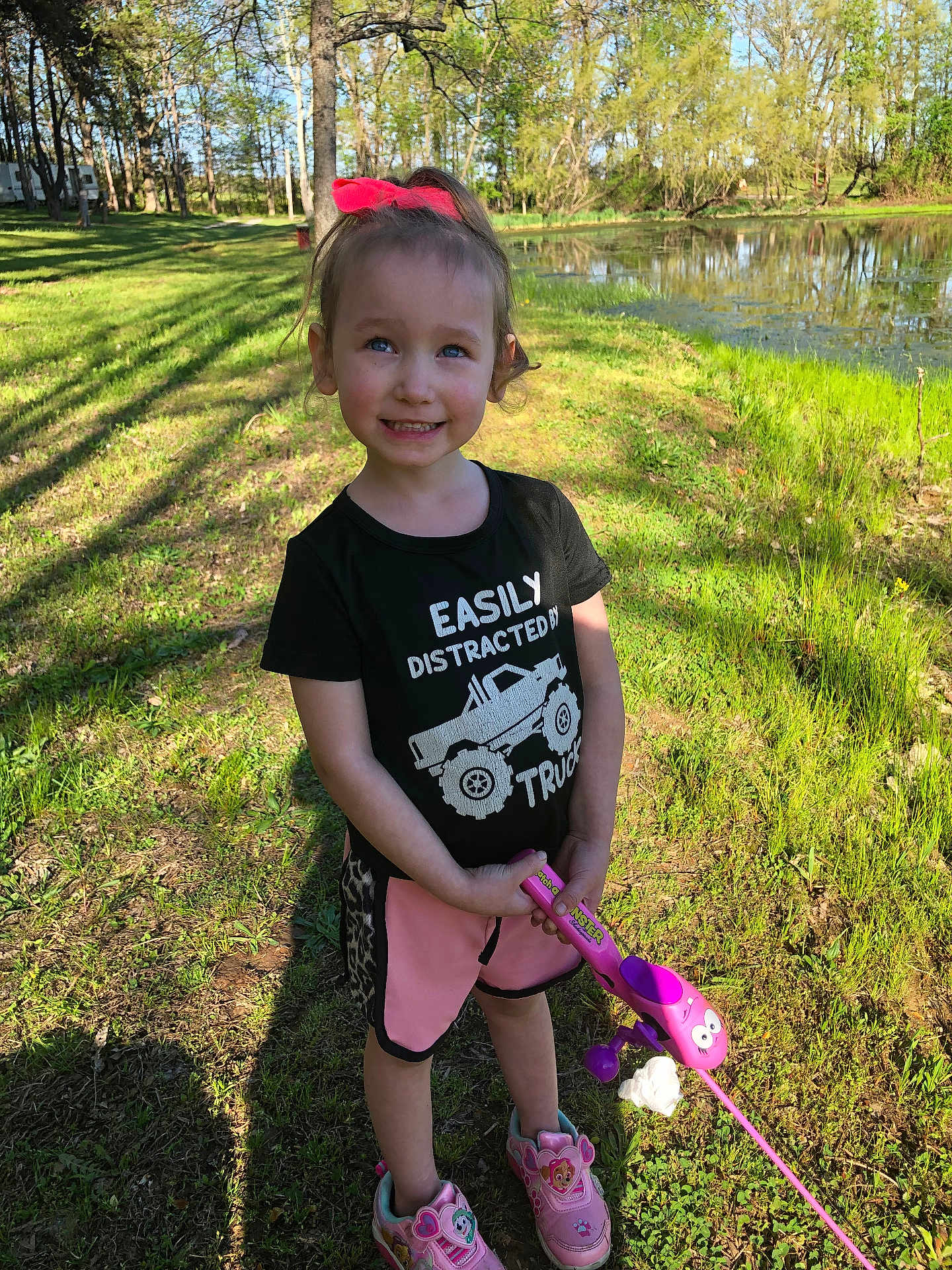 Aubrey is registered to the contest to win money with this photo: child, girl, smiling, outdoor, grass, trees, pond, toy, fishing_rod, red_bow, shorts, black_tshirt, pink_shoes, sunlight, nature, summer, playful, happy, casual_clothing, daytime