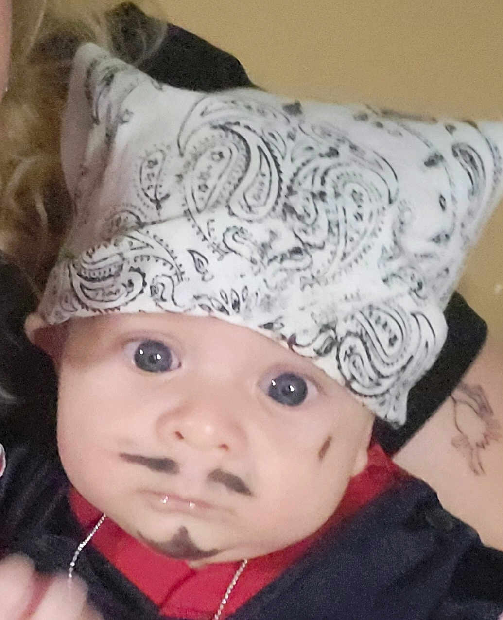 Gaby is registered to the contest to win money with this photo: baby, bandana, face_paint, mustache, goatee, blue_eyes, curly_hair, tattoo, red_clothing, black_clothing, portrait, indoor, person, cute, infant, headwear, expression, closeup, human, child