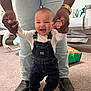 baby, infant, smiling_baby, overalls, denim, adult_hands, supporting, first_steps, boots, jeans, carpet, nursery, crib, toy_bag, indoor, family_moment, happy, portrait, cute, toddler