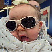 Ella a rejoint le concours — aidez-le/la à gagner de superbes lots ! baby, child, sunglasses, white_jacket, polka_dots, tongue_out, indoor, public_space, person, cute, playful, cheeky, face, headwear, winter_clothing, holding, blurred_background, sign, number, glasses
