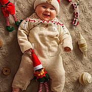 Ella a rejoint le concours — aidez-le/la à gagner de superbes lots ! baby, infant, smiling, knitwear, red_hat, plush_toy, elf_doll, carpet, holiday_decor, christmas_theme, ornaments, cozy, cute, indoors, child, festive, soft_texture, toy, cheerful, winter_clothing