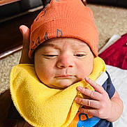 Sargun Kaur joined the competition — help win amazing prizes! baby, infant, orange_hat, yellow_bib, hand, face, indoor, person, clothing, cute, newborn, expression, skin, fingers, holding, carpet, furniture, shirt, closeup, portrait