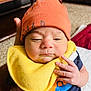 baby, infant, orange_hat, yellow_bib, hand, face, indoor, person, clothing, cute, newborn, expression, skin, fingers, holding, carpet, furniture, shirt, closeup, portrait