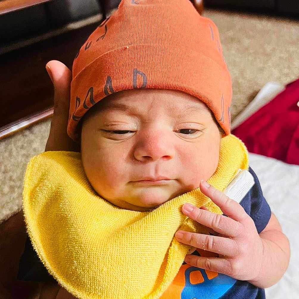 Sargun Kaur joined the competition — help win amazing prizes! baby, carpet, closeup, clothing, cute, expression, face, fingers, furniture, hand, holding, indoor, infant, newborn, orange_hat, person, portrait, shirt, skin, yellow_bib