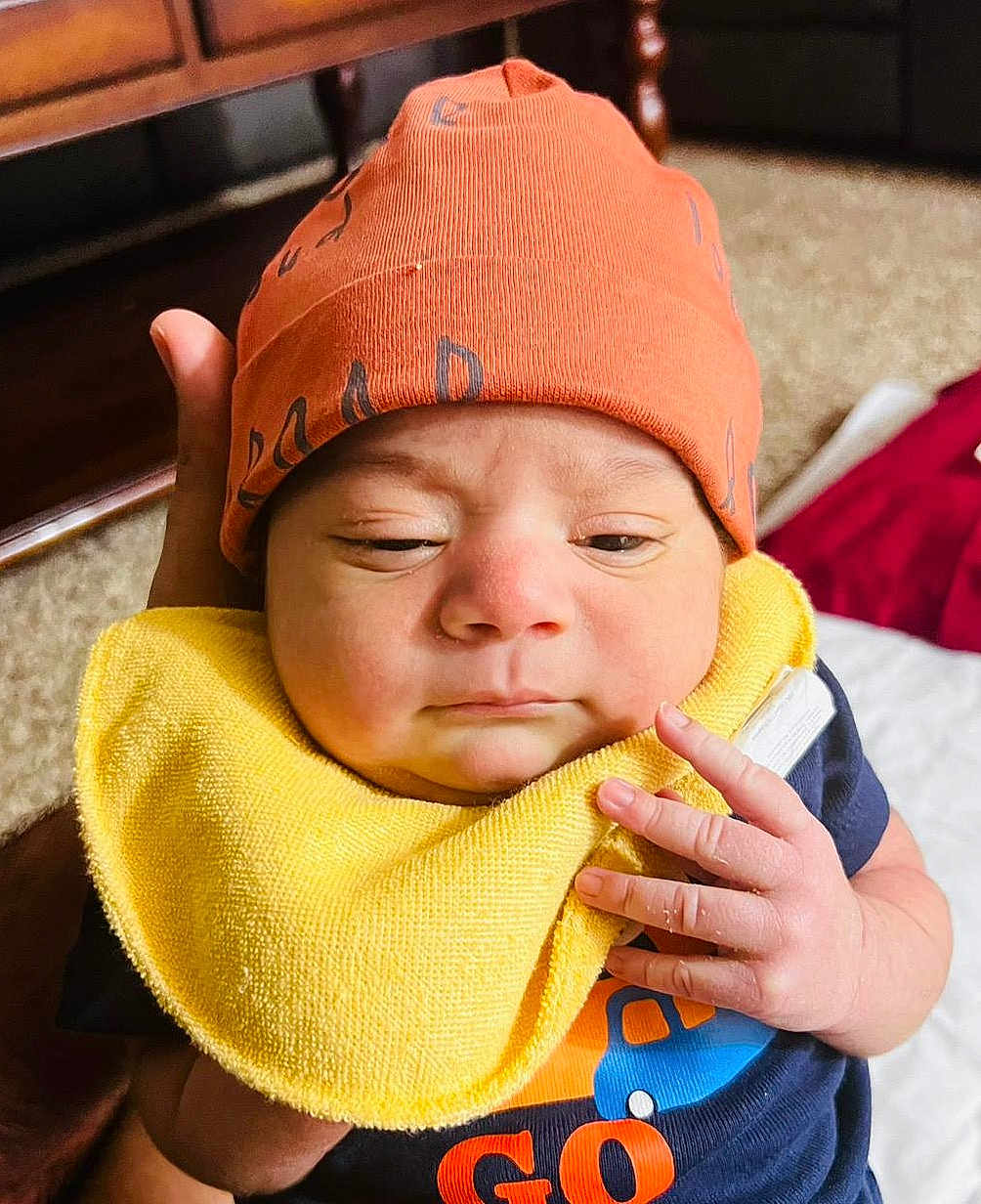 Sargun Kaur joined the competition — help win amazing prizes! baby, infant, orange_hat, yellow_bib, hand, face, indoor, person, clothing, cute, newborn, expression, skin, fingers, holding, carpet, furniture, shirt, closeup, portrait