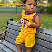 Gael Vulgarin is registered to the contest to win money with this photo: toddler, child, bench, outdoor, park, yellow_clothing, purple_clothing, shorts, sneakers, grass, trees, face, person, curly_hair, holding_food, casual, summer, daytime, expression, portrait