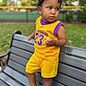toddler, child, bench, outdoor, park, yellow_clothing, purple_clothing, shorts, sneakers, grass, trees, face, person, curly_hair, holding_food, casual, summer, daytime, expression, portrait