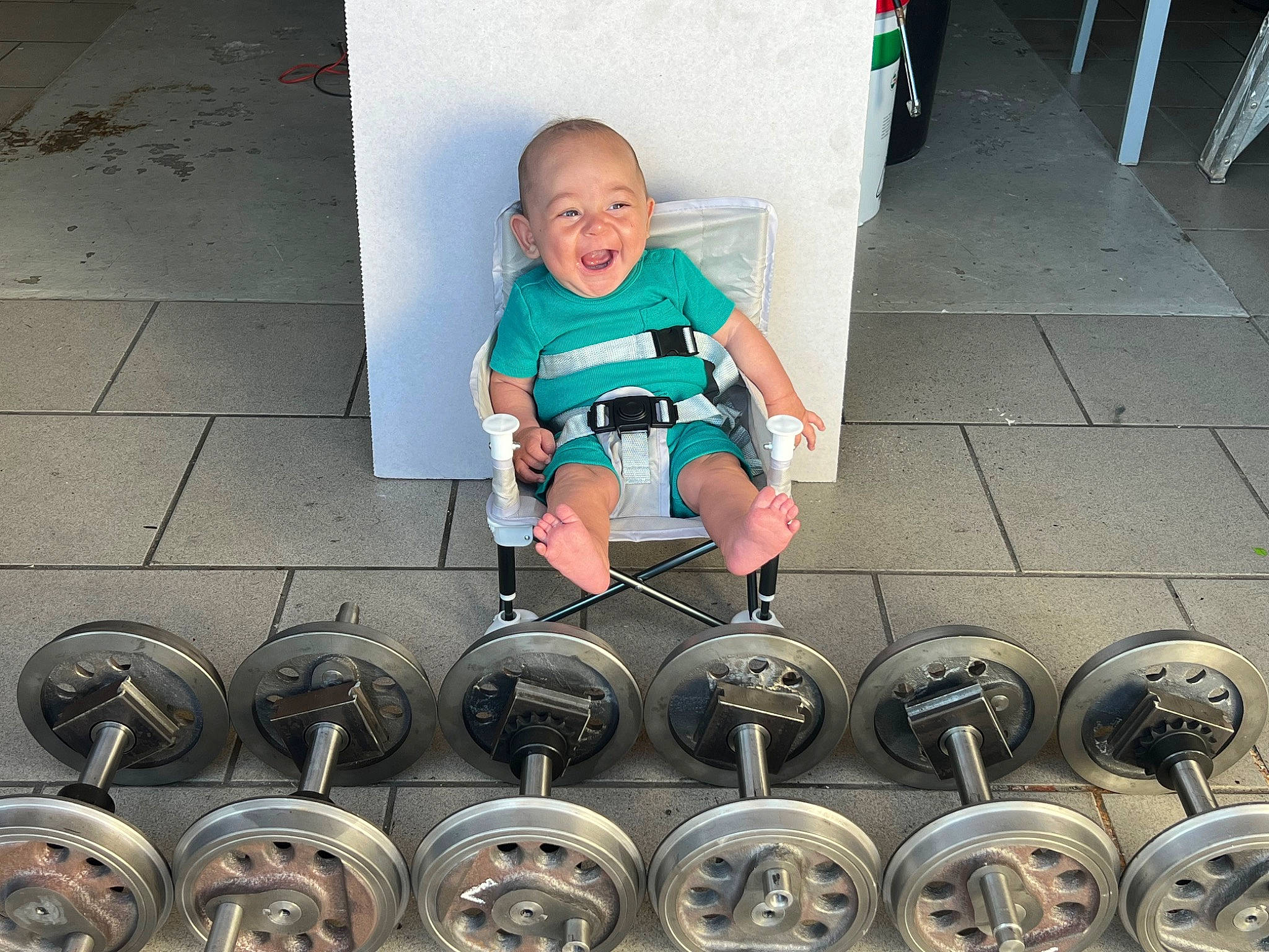 Charlie is registered to the contest to win money with this photo: asphalt, automotive_lighting, automotive_tire, baby, baby_toddler_clothing, electric_blue, flooring, fun, green, motor_vehicle, person, personal_protective_equipment, road, road_surface, rolling, shoe, sidewalk, sitting, smile, sneakers