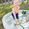 child, boy, smiling, pool, inflatable_pool, water, hose, outdoor, grass, trees, summer, play, happy, shorts, sunlight, backyard, toy, ball, fun, recreation