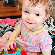 Joyce is registered to the contest to win money with this photo: child, toddler, curly_hair, toy, colorful, indoor, blanket, playful, cute, smiling, clothing, person, face, happy, sitting, floor, hand, eyes, infant, casual