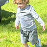 Joyce is registered to the contest to win money with this photo: child, toddler, curly_hair, grass, outdoor, sunlight, sneakers, shorts, long_sleeve_shirt, walking, shadow, nature, person, summer, daylight, greenery, casual_clothing, young, playing, park