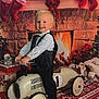 barefoot, child, christmas_stockings, christmas_tree, decorations, festive, fireplace, formal_clothing, gift_boxes, holiday, holiday_decor, indoors, playing, red_rug, smile, smiling, tie, toddler, toy_car, vest