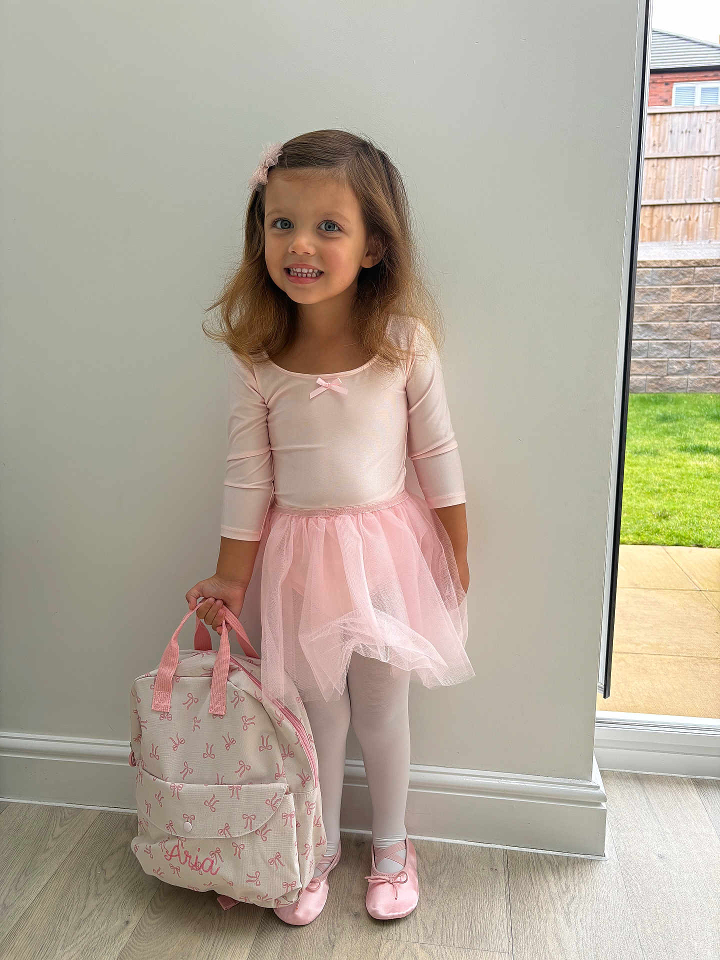 Aria is registered to the contest to win money with this photo: child, girl, ballet, tutu, pink, backpack, indoor, smile, hair_accessory, shoes, tights, floor, window, wall, wood_floor, natural_light, greenery, portrait, standing, cute