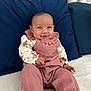 baby, infant, smile, face, clothing, pink_overalls, white_shirt, sitting, cushion, pillow, bed, happy, child, portrait, indoors, cute, young, person, cozy, comfortable