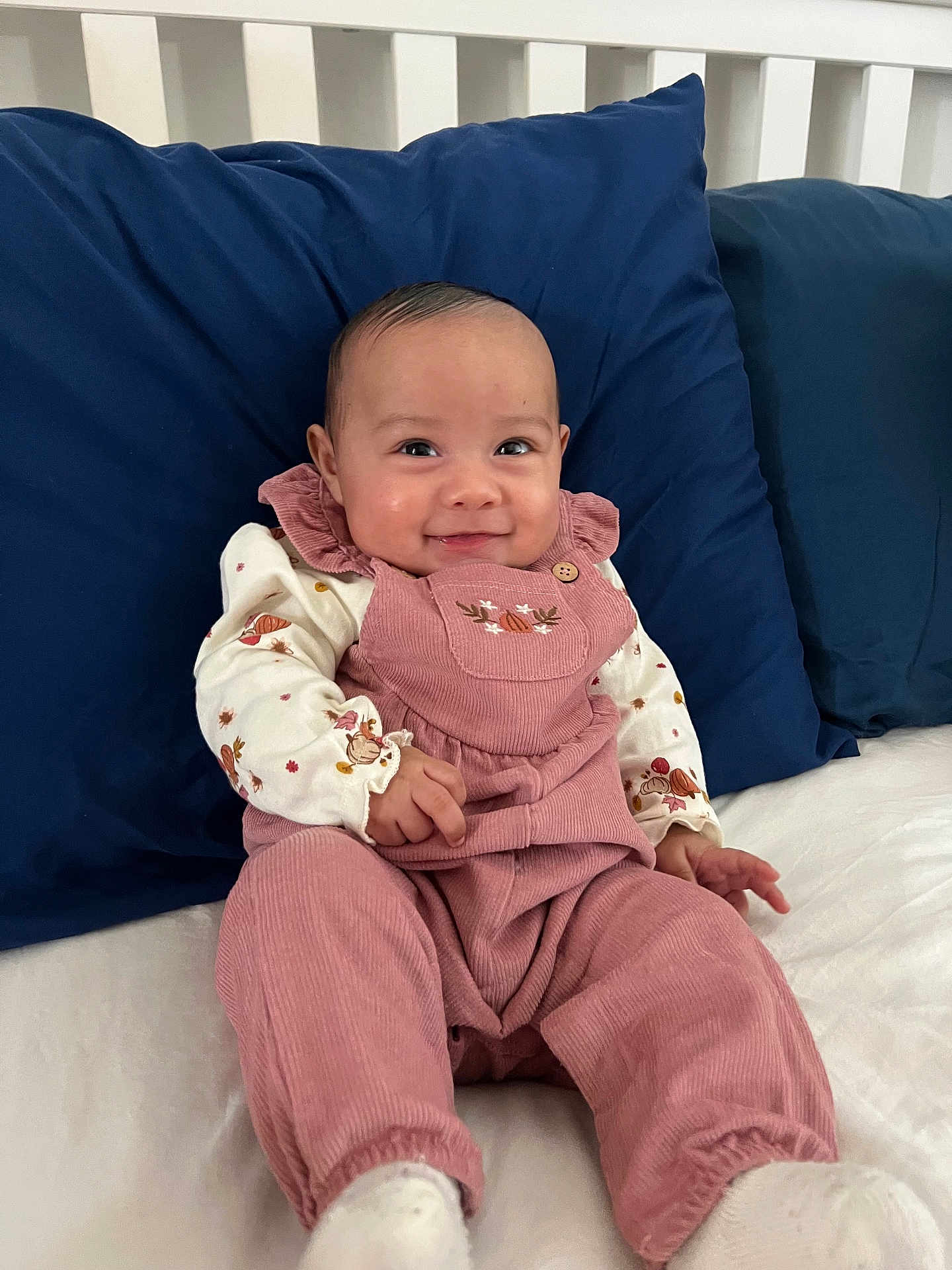 Olivia is registered to the contest to win money with this photo: baby, infant, smile, face, clothing, pink_overalls, white_shirt, sitting, cushion, pillow, bed, happy, child, portrait, indoors, cute, young, person, cozy, comfortable