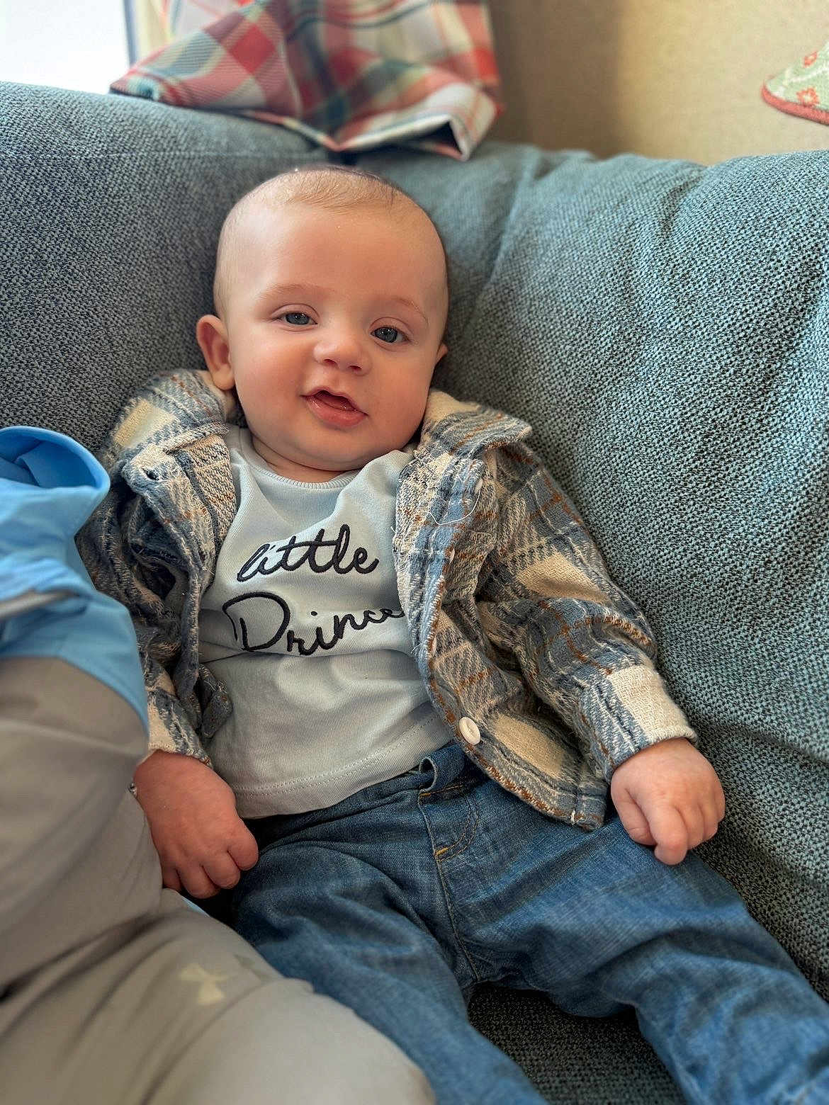 Miles joined the competition — help win amazing prizes! baby, child, plaid_jacket, jeans, couch, blue_couch, smile, relaxed, indoor, portrait, person, clothing, casual_wear, infant, seated, cute, happy, face, furniture, home