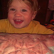 Westley is registered to the contest to win money with this photo: child, toddler, smile, face, blond_hair, yellow_sweater, quilt, furniture, dresser, toy, indoor, happy, person, portrait, head, cheerful, red_eye, clothing, baby, room