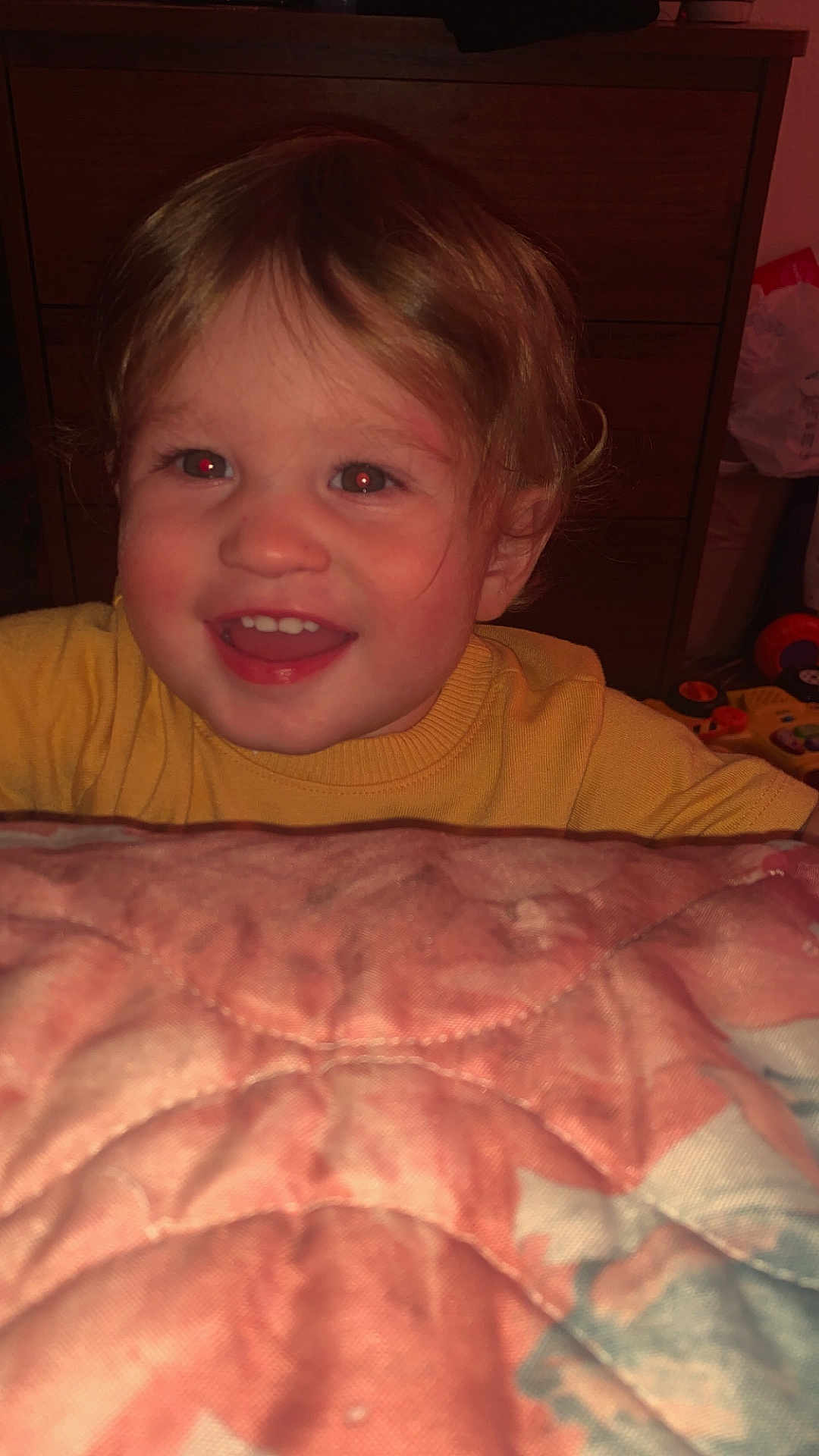 Westley is registered to the contest to win money with this photo: child, toddler, smile, face, blond_hair, yellow_sweater, quilt, furniture, dresser, toy, indoor, happy, person, portrait, head, cheerful, red_eye, clothing, baby, room