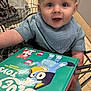Freddie is registered to the contest to win money with this photo: baby, child, shopping_cart, book, bluey, bib, clothing, face, happy, indoor, retail, store, toy, person, seat, cart, floor, smiling, cute, curious