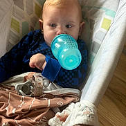 Bradley is registered to the contest to win money with this photo: baby, infant, child, bottle, feeding, pacifier, blue_clothing, blanket, cushion, seated, indoors, portrait, face, eyes, blond_hair, cozy, nursery, wooden_floor, soft_lighting, photography