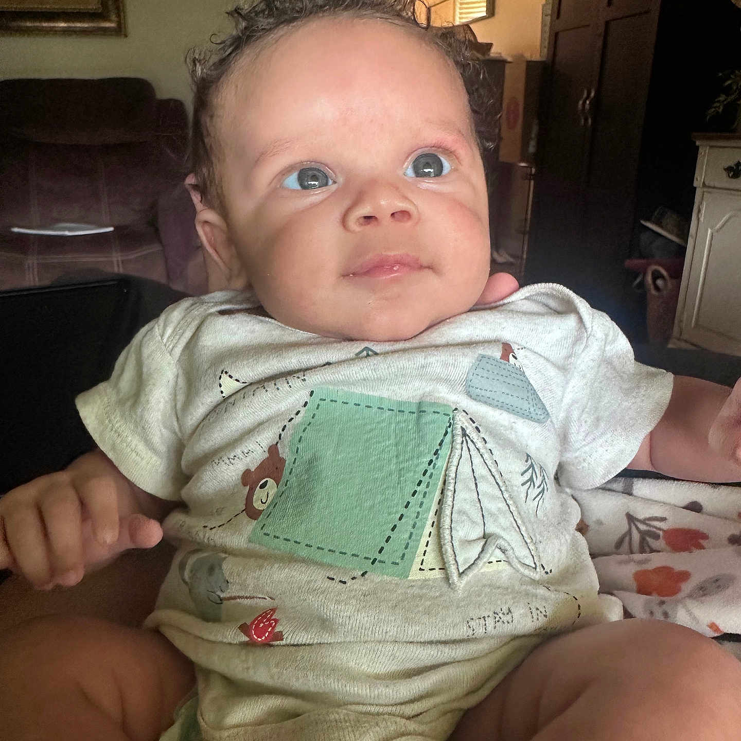 Javien is registered to the contest to win money with this photo: baby, infant, child, onesie, indoor, living_room, curly_hair, cute, person, portrait, sitting, clothing, furniture, couch, hand, face, eyes, skin, expression, young
