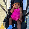 Kylie is registered to the contest to win money with this photo: baby, child, stroller, pink_outfit, blue_socks, outdoor, concrete, curious_expression, infant, headwear, black_headband, seat, legs, feet, person, sunlight, shadow, casual, portrait, one_hand_up