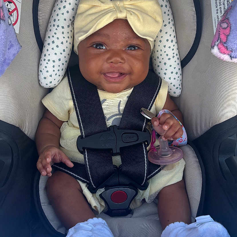 Myla joined the competition — help win amazing prizes! baby, baby_accessory, car_seat, child, closeup, clothing, cute, happy, headband, indoor, infant, pacifier, person, seatbelt, sitting, smiling, socks, straps, toddler, yellow_bow