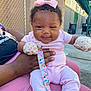 Myla is registered to the contest to win money with this photo: baby, pink_clothing, mittens, bow, pacifier_clip, smiling, curly_hair, outdoor, chain_link_fence, adult_hand, sidewalk, sunlight, seated, happy, infant, person, playful, casual, colorful, daylight