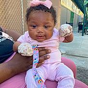 Myla is registered to the contest to win money with this photo: baby, infant, pink_clothing, mittens, bow, pacifier, pacifier_clip, adult_hand, outdoor, chain_link_fence, concrete, sunlight, person, cute, child, holding, sitting, colorful, happy, casual