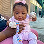 baby, infant, pink_clothing, mittens, bow, pacifier, pacifier_clip, adult_hand, outdoor, chain_link_fence, concrete, sunlight, person, cute, child, holding, sitting, colorful, happy, casual