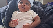 Princeton is registered to the contest to win money with this photo: baby, bear_ears, blanket, blue_hat, casual_clothing, child, couch, cozy, curious, fan, furniture, home, indoor, infant, person, relaxed, soft_texture, striped_pants, television, white_shirt