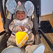 Princeton joined the competition — help win amazing prizes! baby, infant, child, stroller, elephant_costume, gray_clothing, polka_dots, pink_hat, plush_toy, yellow_toy, smiling, tongue_out, sitting, indoors, wooden_floor, black_rug, portrait, front_view, cozy, playful
