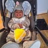 baby, infant, child, stroller, elephant_costume, gray_clothing, polka_dots, pink_hat, plush_toy, yellow_toy, smiling, tongue_out, sitting, indoors, wooden_floor, black_rug, portrait, front_view, cozy, playful
