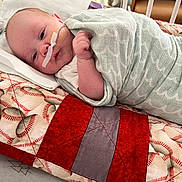 Jace is registered to the contest to win money with this photo: baby, infant, blanket, quilt, hospital, crib, nasal_tube, medical_tape, bed, sleeping, face, person, child, swaddled, soft_fabric, pattern, red, green, closeup, healthcare