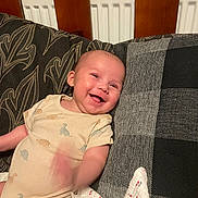 Hunter is registered to the contest to win money with this photo: baby, smile, onesie, dinosaur, cushion, blanket, wooden_headboard, infant, happy, child, indoor, fabric, plaid, pattern, person, cute, face, sitting, cozy, soft