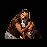 Nala participe au concours pour gagner de l'argent avec cette photo : woman, person, dog, pet, animal, hugging, cuddling, portrait, indoors, black_background, dramatic_lighting, warm_lighting, brown_hair, casual_clothing, white_shirt, shorts, sitting, affection, companion, photography