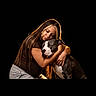 woman, person, dog, pet, animal, hugging, cuddling, portrait, indoors, black_background, dramatic_lighting, warm_lighting, brown_hair, casual_clothing, white_shirt, shorts, sitting, affection, companion, photography
