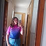 person, mirror, selfie, indoor, jeans, glasses, phone, curly_hair, tshirt, door, hallway, reflection, wood_frame, casual, young_person, scrunchie, sticker, window, tile_floor, curtain