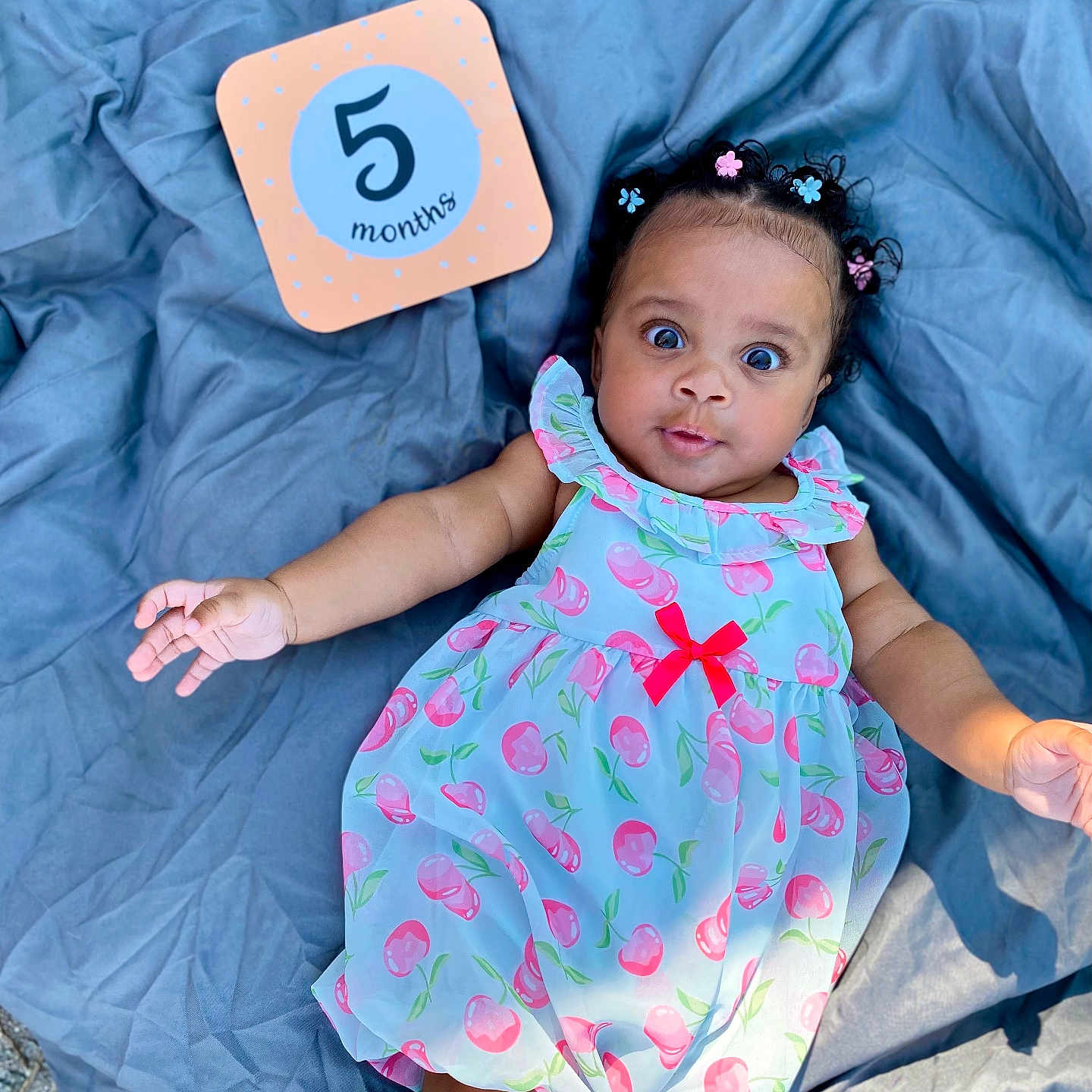 Lolani joined the competition — help win amazing prizes! baby, infant, child, dress, blue_blanket, milestone_card, outdoor, grass, curly_hair, flower_clips, wide_eyes, cute, colorful, portrait, lying_down, summer, skin, smiling, person, pink_cherries