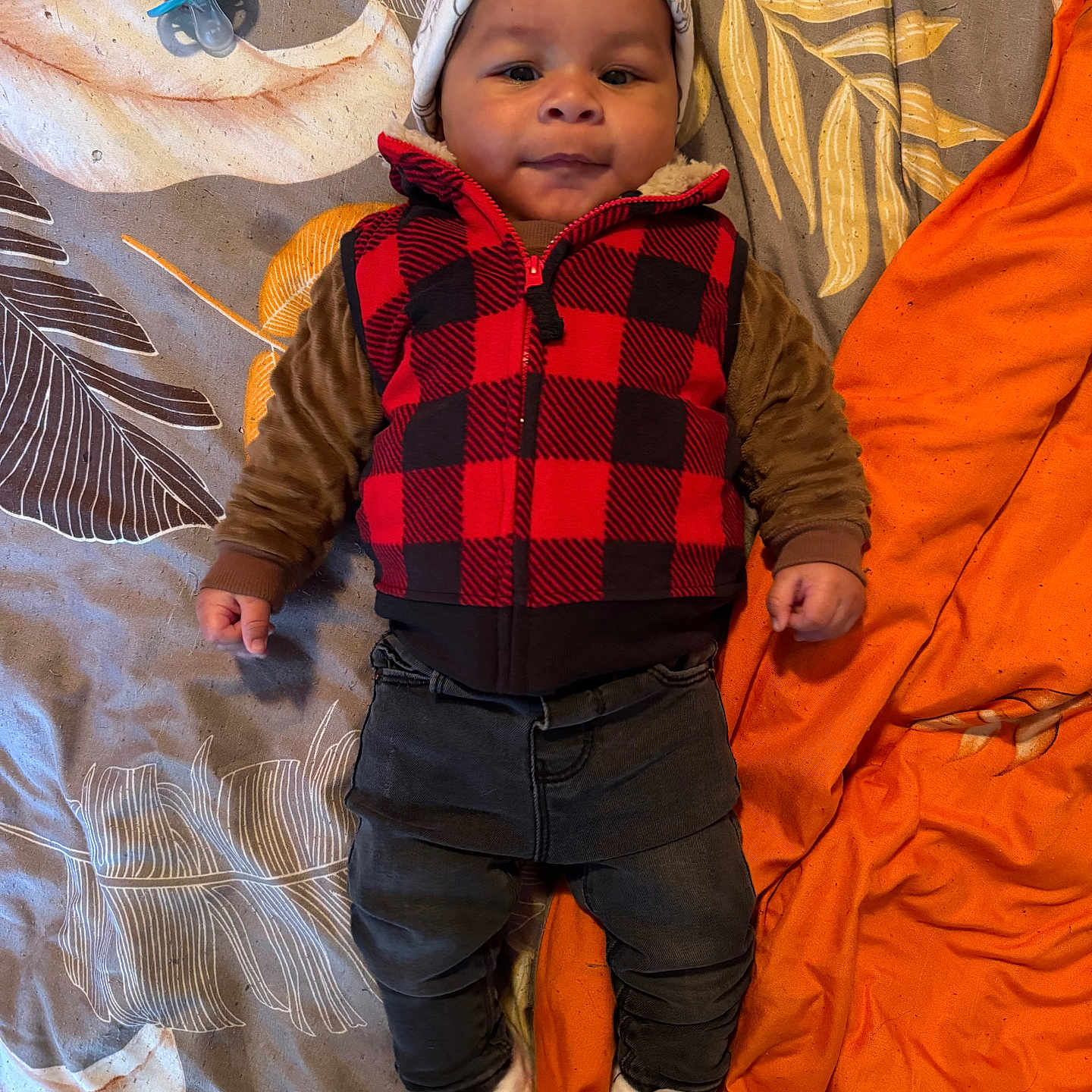 Nikko Lee is registered to the contest to win money with this photo: baby, bedspread, blanket, child, clothing, cute, face, hat, indoor, infant, lying_down, orange, pacifier, pattern, person, portrait, slippers, smiling, vest, warm_clothes