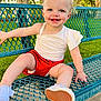 bench, blue_eyes, casual_clothing, child, cute, grass, greenery, happy, metal_bench, outdoor, park, red_bow, red_shorts, shoe, sitting, smile, sunlight, toddler, tree, white_shirt