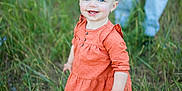 Nikkole is registered to the contest to win money with this photo: adult_legs, blue_eyes, casual_clothing, child, curly_hair, cute, daylight, field, grass, happy, nature, orange_dress, outdoor, person, portrait, smiling, standing, toddler, white_shoes, young_child