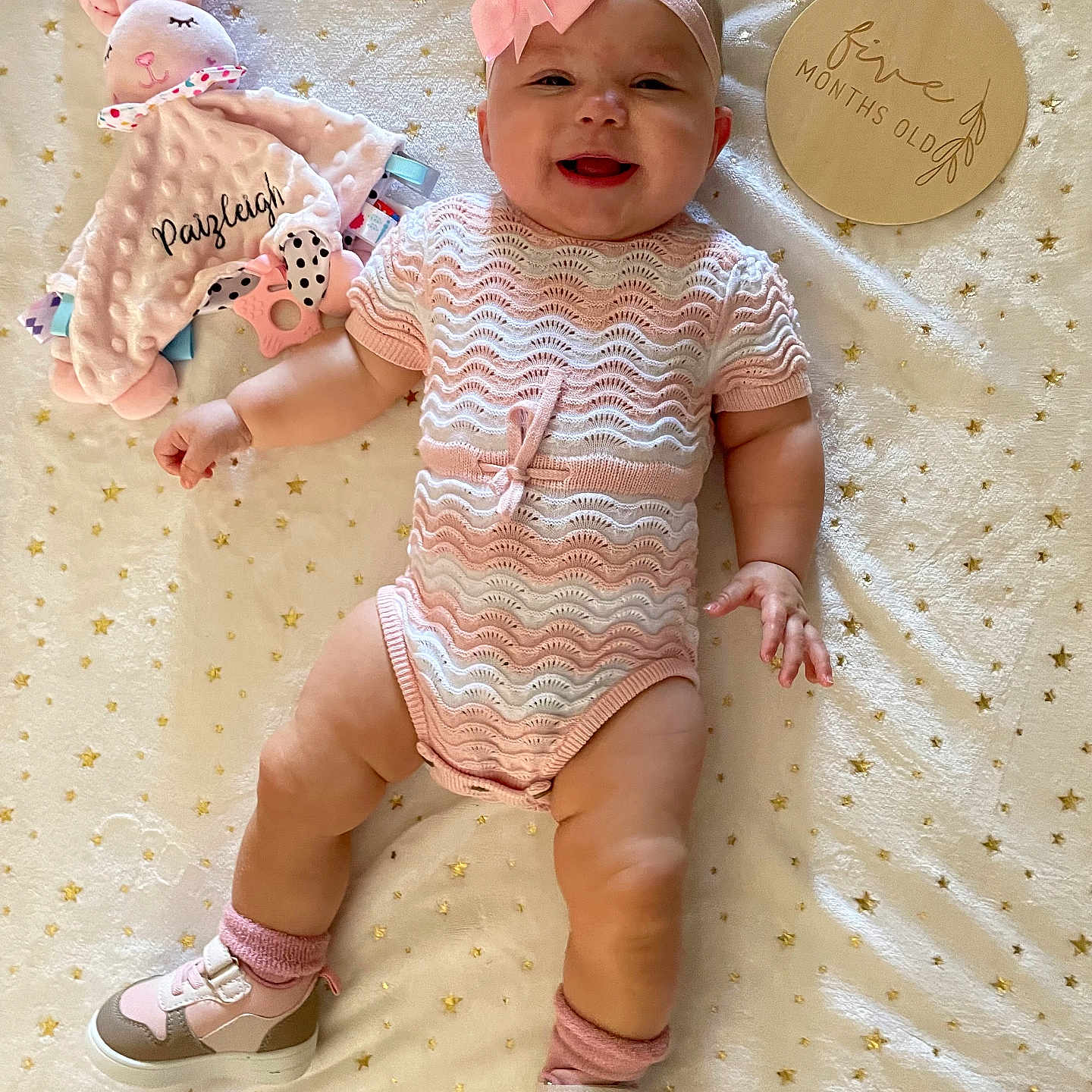 Paizleigh is registered to the contest to win money with this photo: baby, blanket, bow, cute, girl, happy, headband, indoor, infant, knitted_clothing, lying_down, person, plaque, shoes, smile, socks, star_pattern, stuffed_animal, toy, wooden_sign