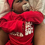 Jahmir is registered to the contest to win money with this photo: baby, pacifier, red_clothing, beanie, infant, blanket, adult_arm, sleepwear, indoor, portrait, child, cute, person, expression, closeup, cozy, resting, wearing, sitting, comfort