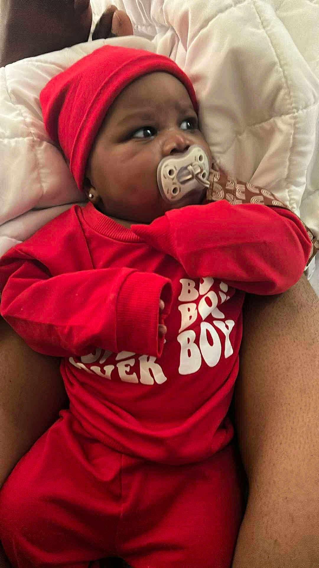 Jahmir is registered to the contest to win money with this photo: baby, pacifier, red_clothing, beanie, infant, blanket, adult_arm, sleepwear, indoor, portrait, child, cute, person, expression, closeup, cozy, resting, wearing, sitting, comfort