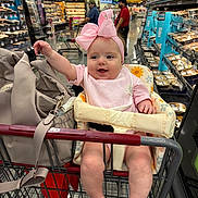 Koa joined the competition — help win amazing prizes! baby, infant, shopping_cart, supermarket, headband, pink_bow, bib, baby_seat, purse, packaged_food, grocery_aisle, smiling, bare_feet, legs, adult_in_background, store_signage, metal_cart, fluorescent_lighting, candid_portrait, family_shopping